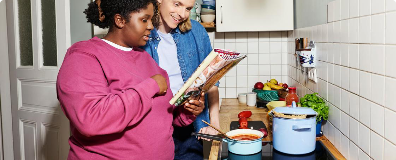 Zwei Personen in einer Kueche, eine haelt eine Kochzeitschrift, die andere kocht